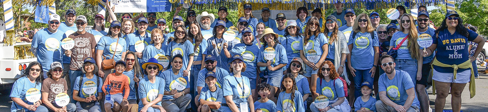 A large group of volunteers and staff members wearing Give Day shirts pose in front of the One Aggie Network and Give Day parade tram.
